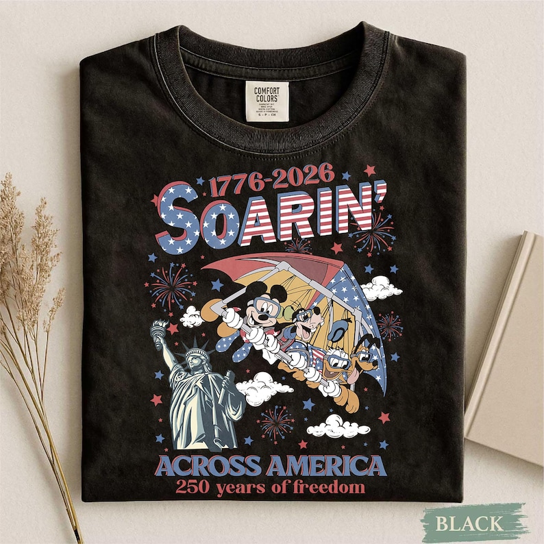Może przedstawiać: Czarny t-shirt z grafiką Myszki Miki, Kaczora Donalda i Goofy'ego szybujących na paralotni nad Statuą Wolności. Koszulka zawiera napis "1776-2026 Soarin' Across America 250 years of freedom" w kolorach czerwonym, białym i niebieskim.