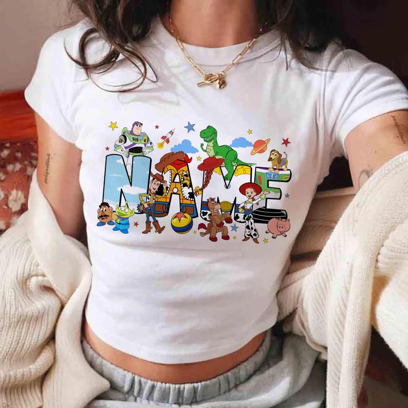 May include: White t-shirt featuring a colorful graphic design with the word "NAME" in large, stylized letters. The design incorporates various cartoon characters and elements, including a rocket, stars, and a dinosaur.