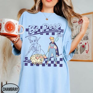 K&ouml;nnte beinhalten: Ein hellblaues T-Shirt mit einem Grafikdruck der Figur Bo Peep und drei Schafen. Das Design enth&auml;lt den Text Bo Peep 199s, einen violetten karierten Hintergrund sowie eine stilisierte Strichzeichnung der Figur neben einer farbigen Illustration.