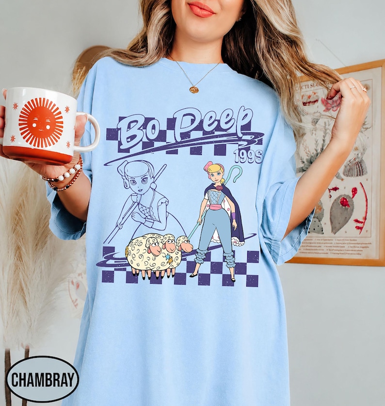 K&ouml;nnte beinhalten: Ein hellblaues T-Shirt mit einem Grafikdruck der Figur Bo Peep und drei Schafen. Das Design enth&auml;lt den Text Bo Peep 199s, einen violetten karierten Hintergrund sowie eine stilisierte Strichzeichnung der Figur neben einer farbigen Illustration.
