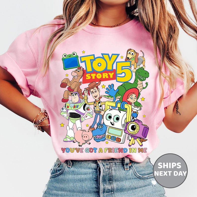 May include: A light pink t-shirt featuring a colorful graphic of Toy Story characters including Woody, Buzz Lightyear, Rex, and Slinky Dog. The design includes the text TOY STORY 5 and YOU'VE GOT A FRIEND IN ME in multi-colored lettering.