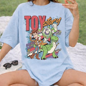May include: Light blue t-shirt featuring a graphic of Woody, Buzz Lightyear, and Rex from Toy Story. The design includes the text "TOY STORY" in red and orange, with "Since 1995" and character names. The shirt has short sleeves.