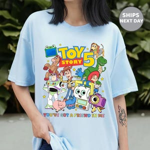 May include: A light blue t-shirt featuring a colorful graphic of Toy Story characters including Woody, Buzz Lightyear, Jessie, Rex, Slinky Dog, and others. The text reads "TOY STORY 5" and "YOU'VE GOT A FRIEND IN ME" in bright, multi-colored lettering.