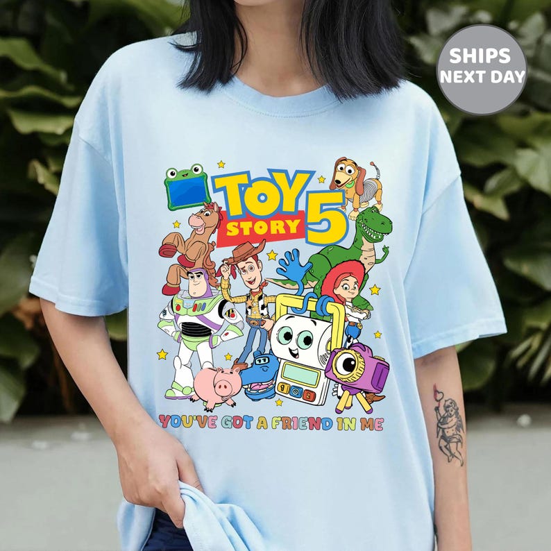 May include: A light blue t-shirt featuring a colorful graphic of Toy Story characters including Woody, Buzz Lightyear, Jessie, Rex, Slinky Dog, and others. The text reads "TOY STORY 5" and "YOU'VE GOT A FRIEND IN ME" in bright, multi-colored lettering.