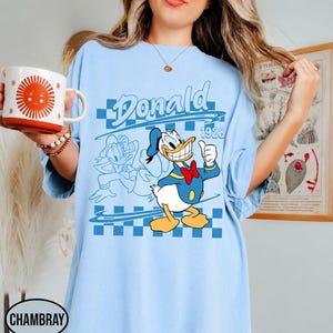 May include: A light blue t-shirt featuring a graphic of Donald Duck with the text "Donald" and "1928" over a blue checkered pattern. The design includes a full-color illustration of the character alongside a blue line-art outline of the same figure.
