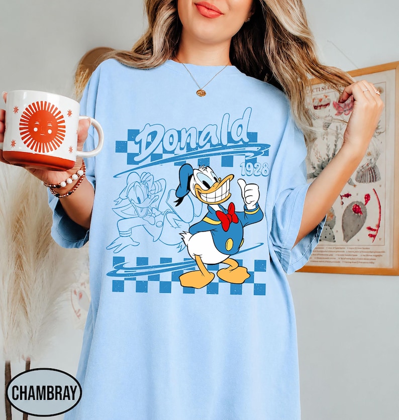May include: A light blue t-shirt featuring a graphic of Donald Duck with the text "Donald" and "1928" over a blue checkered pattern. The design includes a full-color illustration of the character alongside a blue line-art outline of the same figure.