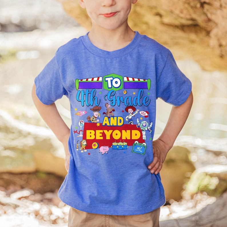 May include: A blue short-sleeved t-shirt featuring a graphic print with the text TO 4th Grade AND BEYOND. The design includes colorful illustrations of various animated toy characters, including a cowboy, a cowgirl, a space ranger, and others.