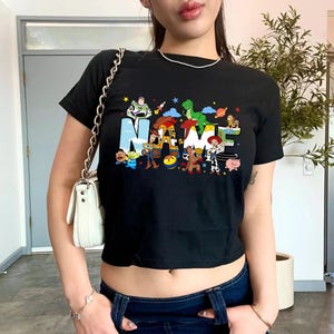 May include: Black crop top featuring the word "NAME" in a colorful, cartoonish font, with various character illustrations. The shirt is short-sleeved and has a crew neck.