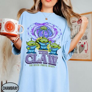 May include: A light blue t-shirt featuring a graphic of three green three-eyed aliens in blue space suits standing beneath a purple swirling portal and a mechanical claw. The text reads THE CLAW and THE MYSTIC PORTAL AWAITS in purple and green.