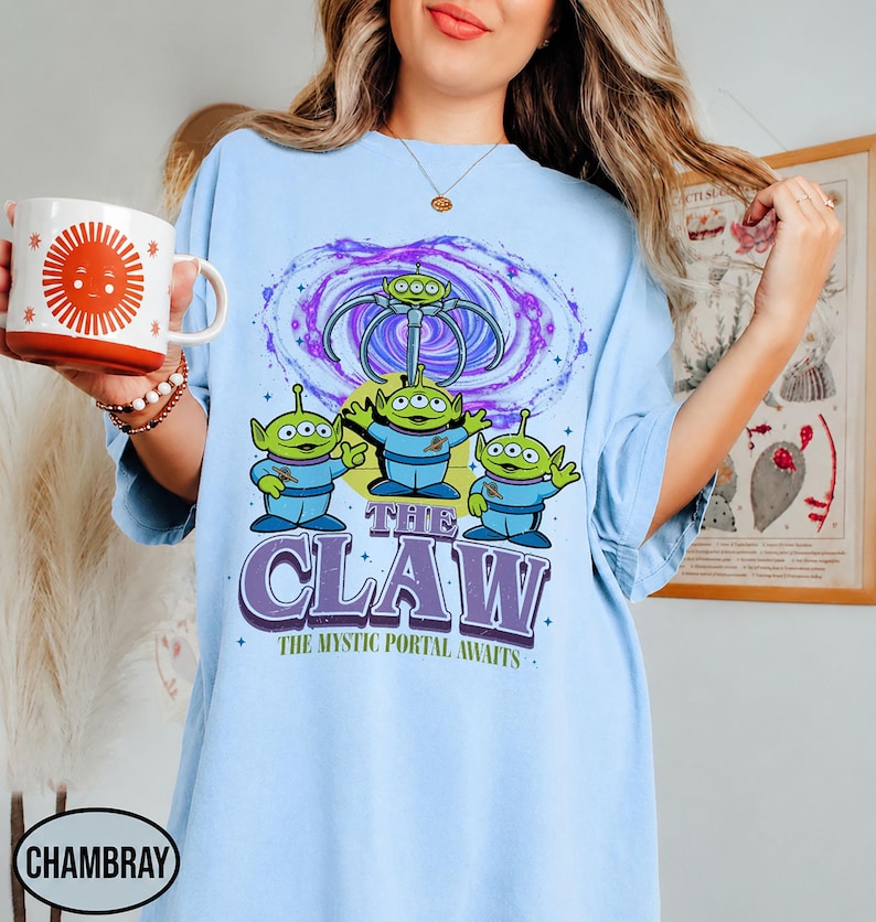 May include: A light blue t-shirt featuring a graphic of three green three-eyed aliens in blue space suits standing beneath a purple swirling portal and a mechanical claw. The text reads THE CLAW and THE MYSTIC PORTAL AWAITS in purple and green.