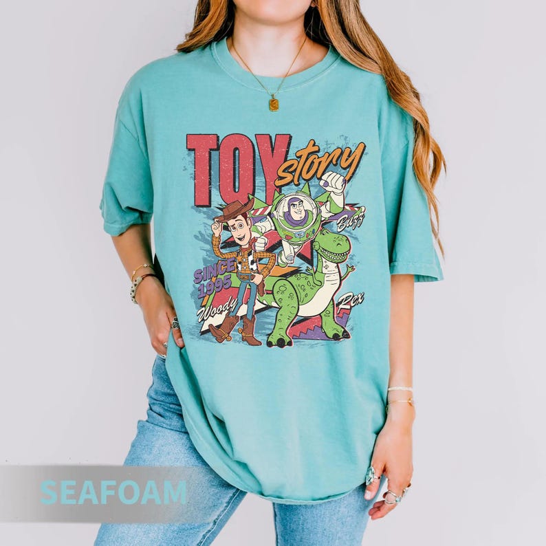 May include: Seafoam green t-shirt featuring the words "Toy Story" in red and yellow, with Woody, Buzz Lightyear, and Rex the dinosaur. The shirt has the text "Since 1995" and is a relaxed fit.