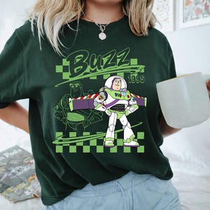 K&ouml;nnte beinhalten: Ein dunkelgr&uuml;nes T-Shirt mit einer Grafik von Buzz Lightyear und dem Text "BUZZ" sowie "199S" &uuml;ber und neben der Figur. Das Design enth&auml;lt einen hellgr&uuml;nen Hintergrund mit Schachbrettmuster und eine Strichzeichnung der Figur dahinter.