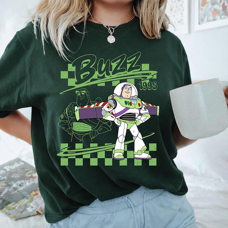 K&ouml;nnte beinhalten: Ein dunkelgr&uuml;nes T-Shirt mit einer Grafik von Buzz Lightyear und dem Text "BUZZ" sowie "199S" &uuml;ber und neben der Figur. Das Design enth&auml;lt einen hellgr&uuml;nen Hintergrund mit Schachbrettmuster und eine Strichzeichnung der Figur dahinter.