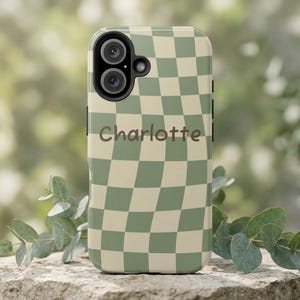 May include: A phone case with a green and cream checkered pattern. The case has a camera cutout and the name "Charlotte" printed in a script font. The case is shown on a stone surface with green foliage in the background.