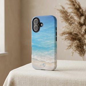 May include: A phone case featuring a serene beach scene with blue ocean waves and a sandy shore. The case has a blue and white gradient design, with the initials "AG" in a delicate script. The case is designed for a smartphone.