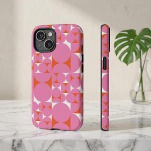 May include: A pink and orange geometric pattern phone case. The case features a repeating design of circles and quarter-circles in pink, orange, and white. The phone case is shown from the front and side, showcasing the design and the phone's camera.