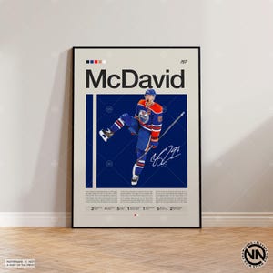 以下が含まれることがあります： 青とオレンジのジャージを着たホッケー選手とMcDavidの文字が描かれた縦型の額入りポスター。デザインには、濃い青の中央パネル、白いサイン、キャリアの統計や功績を詳述した小さなテキストブロックが含まれています。