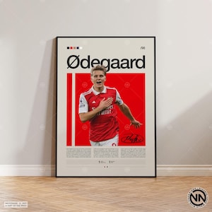 May include: A framed poster featuring a photograph of a soccer player in a red and white jersey against a red background. The text reads Ødegaard at the top, includes a signature, and contains biographical information in small black text below.