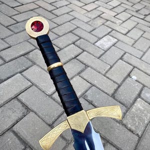 May include: A sword featuring a metallic gold crossguard and pommel with intricate etched patterns. The pommel is set with a large, faceted red gemstone. The handle is wrapped in black material, accented by a central gold ring near the grip.