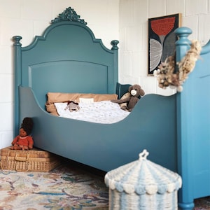 Könnte beinhalten: Ein teal-farbener Bettrahmen mit dekorativem Kopfteil. Das Bett ist mit Kissen, einer Decke und Stofftieren ausgestattet. Ein Weidenkorb und ein gestreifter Korb befinden sich im Vordergrund. Ein farbenfroher Teppich und Kunstwerke vervollständigen die Raumdekoration.