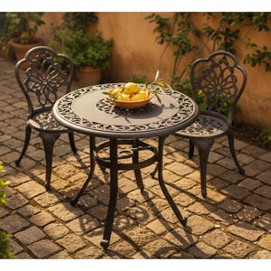 May include: A black metal patio set with a round table and two chairs. The table has a decorative floral design on the tabletop and a matching design on the chair backs. A wooden bowl of lemons sits on the table.