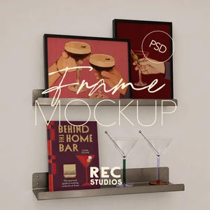 May include: Two horizontal metal wall shelves holding two framed art prints, a book titled Behind the Home Bar by Cara Devine, and two martini glasses with colored stems. The top frames display illustrations of hands holding cocktails.