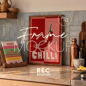Può includere: Una cornice in legno contenente un poster con sfondo rosso e rosa, un'illustrazione di un peperoncino rosso con gambo verde e il testo bianco CHILLI. Accanto si trovano un libro di cucina Pasta Grannies Eat Everything, una bottiglia in rame e una ciotola.