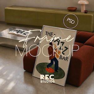 May include: A silver-framed poster mockup featuring a graphic illustration of a musician playing a saxophone on a vinyl record. The text on the poster reads THE JAZZ BAR. The frame is positioned on the floor in front of a rust-colored armchair.