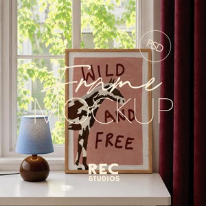 Puede incluir: Un marco de fotos de madera clara que contiene una impresión de un caballo con manchas marrones y blancas sobre un fondo rosa polvoriento. El texto WILD AND FREE está impreso en marrón oscuro. Una lámpara azul con base redonda está a la izquierda.