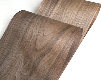 May include: A flexible sheet of brown walnut wood veneer featuring a natural grain pattern with visible knots and swirls. The thin material is curved into two overlapping loops, showcasing the smooth texture and rich wood tones of the surface.