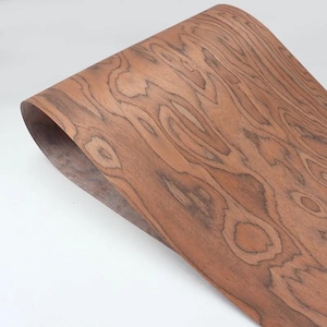 Puede incluir: Una lámina de chapa de madera de color marrón rojizo intenso, con un patrón de veta pronunciado. La chapa está curvada, mostrando la flexibilidad del material. La veta presenta patrones arremolinados y vetas más oscuras.