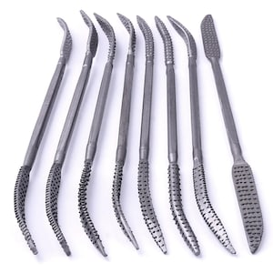 May include: A set of eight grey metal sculpting tools, each featuring a long, slender handle and textured, abrasive rasp-like ends. The tools vary in shape, with some having curved, pointed tips and others featuring flat, rectangular ends for shaping.
