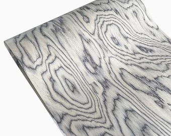May include: Close-up of a curved wood veneer. The wood has a light grey base with dark grey swirling lines and patterns. The natural grain creates a textured appearance. The wood is curved.