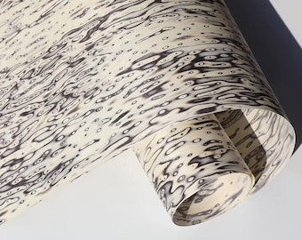 May include: A rolled sheet of paper featuring a cream-coloured backdrop with a pattern of dark grey, organic shapes, reminiscent of water ripples. The design incorporates small, dark grey dots dispersed throughout.