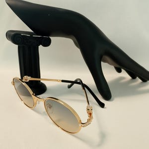 May include: Gold-tone oval sunglasses featuring thin metal frames with crystal-encrusted panther head accents at the hinges. The lenses have a light gradient tint, and the temple tips are finished in black. The design includes adjustable nose pads.