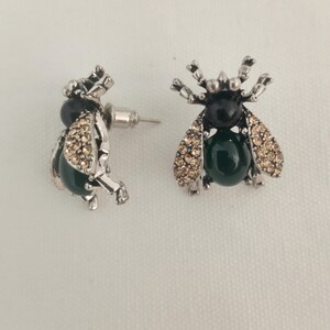 May include: A pair of silver-toned insect-shaped stud earrings featuring a black cabochon head, a dark green oval cabochon body, and wings encrusted with small champagne-colored crystals. The earrings have a post-back closure mechanism.