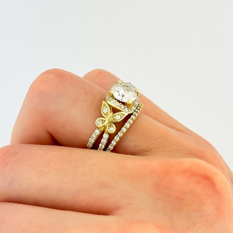May include: A gold ring set with a large, round, clear diamond. The ring is accented with two butterfly designs on either side of the diamond. The ring is worn on a person's finger.