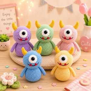 May include: Five small, round, crocheted monster dolls in purple, green, pink, blue, and yellow. Each creature features two yellow horns, a single large black eye with a white border, and a smiling mouth with two small red teeth. The dolls are arranged on a round, woven mat.