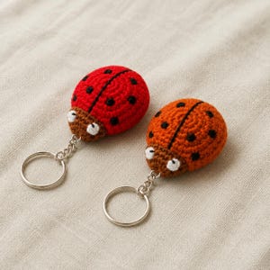 Pode incluir: Dois porta-chaves de joaninha em croché com argolas de metal prateado. Uma joaninha é vermelho-brilhante com pintas pretas e a outra é laranja com pintas pretas. Ambas têm cabeças castanhas com pequenos olhos brancos e uma corrente prateada.