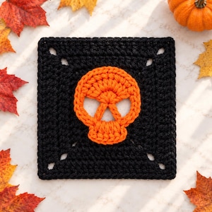 May include: A square crochet granny square featuring a solid black background with a centered, bright orange skull motif. The skull design is created with thick yarn stitches, showcasing open eye sockets and a defined jawline in a flat, square format.