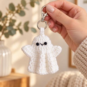 May include: A white crocheted ghost keychain featuring two small black bead eyes and a stitched black mouth. The ghost is attached to a silver-toned metal lobster clasp and a circular split ring, allowing it to be easily clipped onto bags or keys.