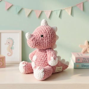 Peut inclure: Une peluche dinosaure rose crochetée avec des détails blancs sur les cornes, les épines et les pieds. Le jouet en peluche a des taches blanches et une petite étiquette "Crochet Critters". Le dinosaure est sur une surface blanche.