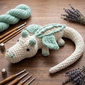 May include: A crocheted dragon toy in cream and mint green yarn with black bead eyes, accompanied by a skein of mint green yarn, several wooden-handled crochet hooks, two metal thimbles, and sprigs of dried lavender arranged around it.