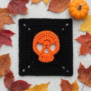 May include: A square crochet motif featuring a black background with a centred orange skull design. The piece is crafted with visible yarn stitches, creating a flat, geometric shape with openwork corners, showcasing a distinct contrast between the colours.