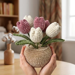 May include: A crochet tulip bouquet featuring soft velvet-textured flowers in shades of dusty rose and cream. The tulips have deep green crocheted leaves and are arranged in a round, textured beige crocheted pot with a visible stitch pattern.