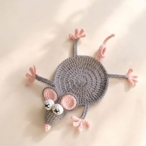 Könnte beinhalten: Ein handgefertigter Untersetzer in Form einer Häkelmaus. Der Untersetzer ist hauptsächlich grau mit rosa Akzenten an Ohren, Füßen und Schwanz. Die Maus hat weiße Augen mit schwarzen Kreuzdetails.