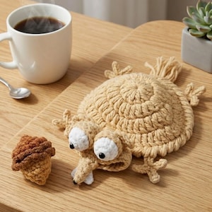 May include: A whimsical, beige crocheted frog-shaped coaster with large white and black eyes, and a small crocheted acorn. A white mug of coffee and a succulent plant in a gray pot are in the background. The items are on a light wood surface.