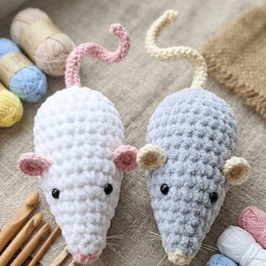 May include: Two plush crochet mice, one white with pink ears and tail, the other light gray with cream ears and tail. Both feature black bead eyes, small noses, and thin whiskers. The mice are crafted from soft, textured yarn and are displayed alongside wooden crochet hooks and various colorful yarn skeins.