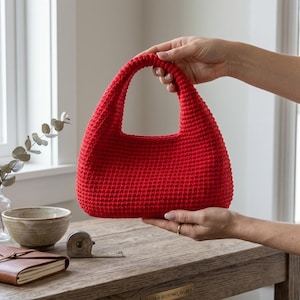Puede incluir: Un bolso de ganchillo de color rojo vibrante con una silueta estructurada y redondeada y un asa única integrada. El bolso está elaborado con un patrón de puntos denso y uniforme, creando una superficie texturizada. El diseño es sencillo y compacto.