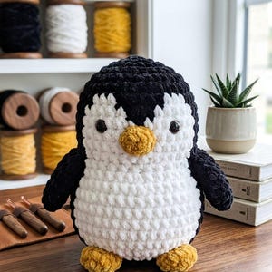 May include: A plush penguin toy made of soft, chunky yarn with a black head and back, a white belly, and a yellow beak and feet. The penguin has two dark, round eyes and is positioned on a wooden surface with crochet hooks and yarn spools.
